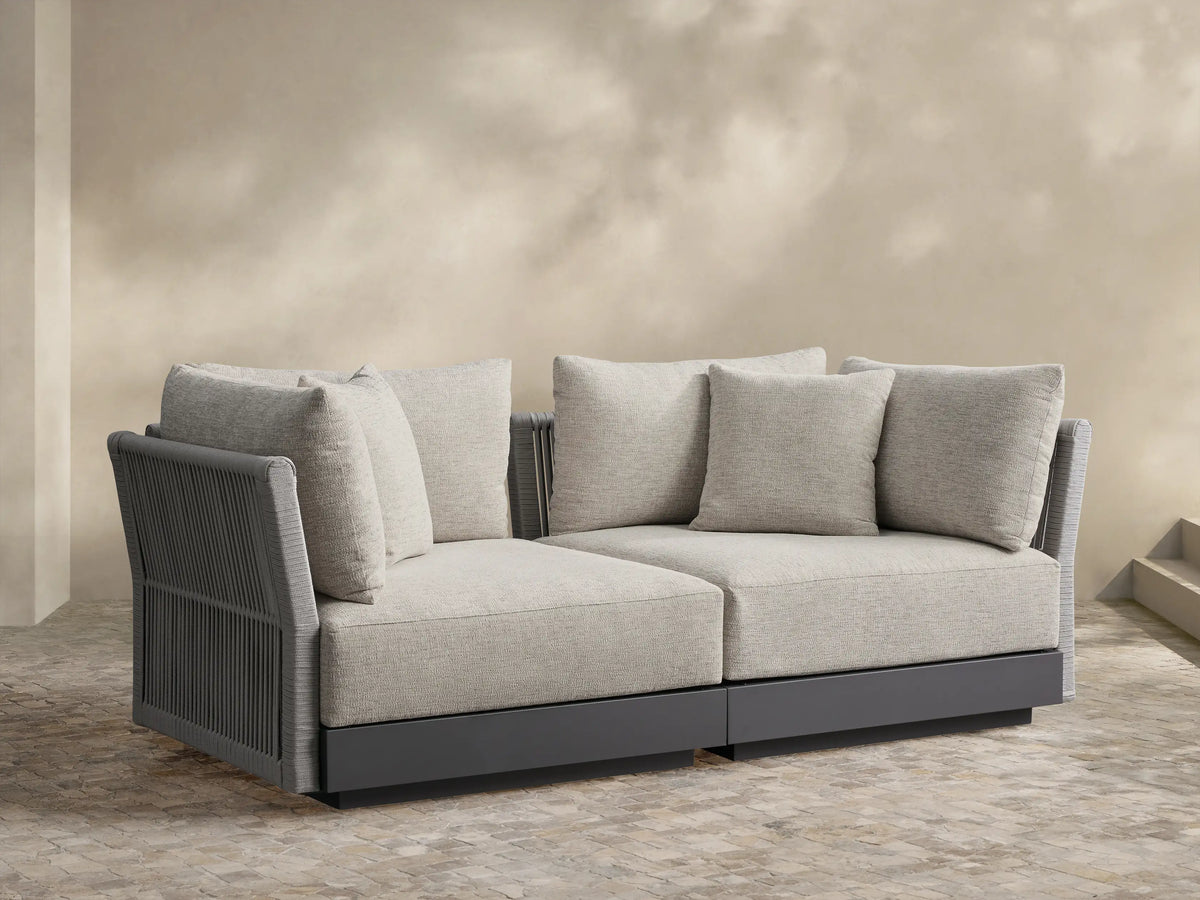 Naxos Two-Piece Modular Sofa with light gray textured cushions, dark gray frame, and ribbed arms on a stone patio.