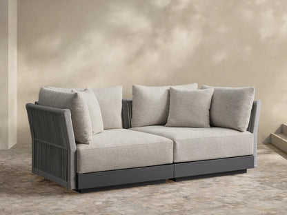 Naxos Two-Piece Modular Sofa with light gray textured cushions, dark gray frame, and ribbed arms on a stone patio.