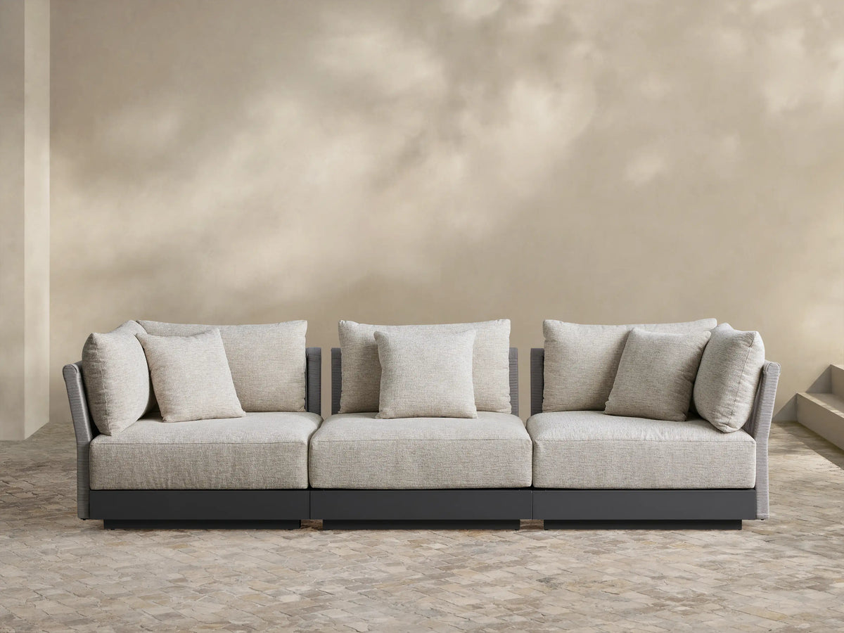 Naxos 3-piece modular sofa: oatmeal cushions, dark gray frame with woven arms, on stone patio against beige wall.
