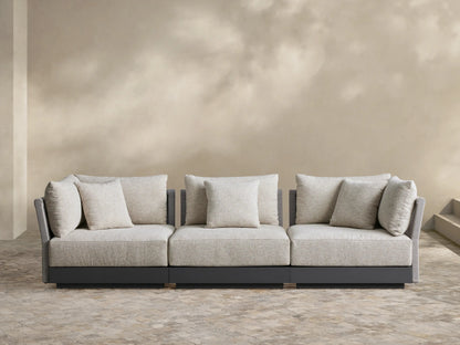 Naxos 3-piece modular sofa: oatmeal cushions, dark gray frame with woven arms, on stone patio against beige wall.