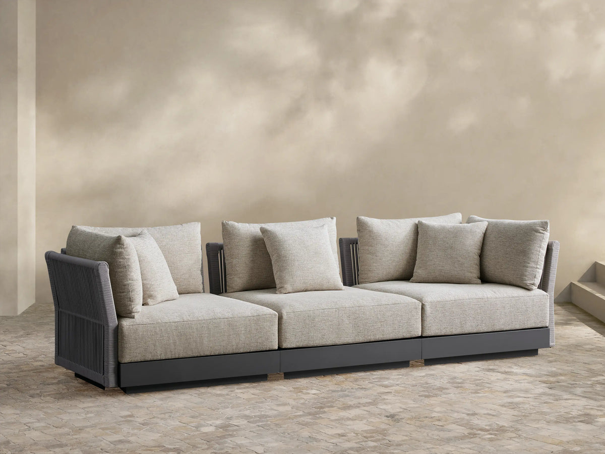 Naxos 3-piece modular sofa: oatmeal cushions, dark gray frame with woven arms, on stone patio against beige wall.