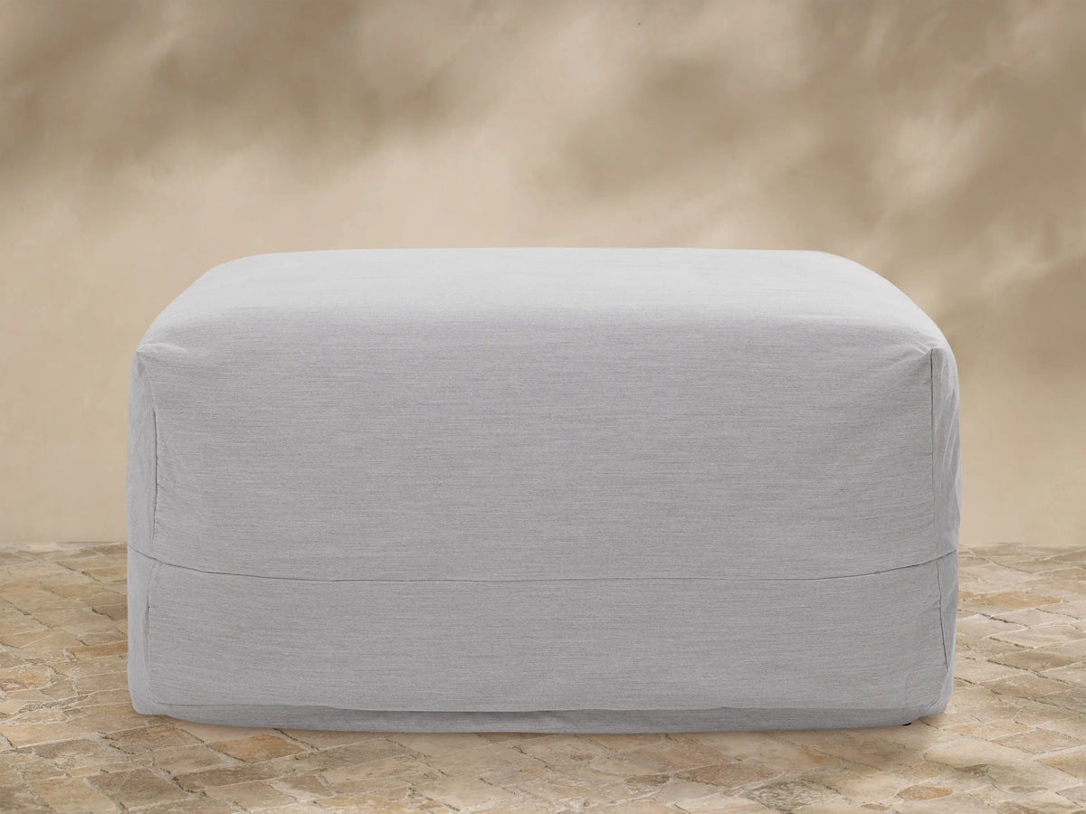 Light gray Naxos Outdoor Collection Covers on a sofa, lounge chair, and table on a stone patio with plants and a tan wall.