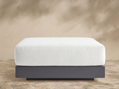 Naxos Outdoor Ottoman: rectangular white cushion on a dark gray aluminum base, centered on a light-colored patio.