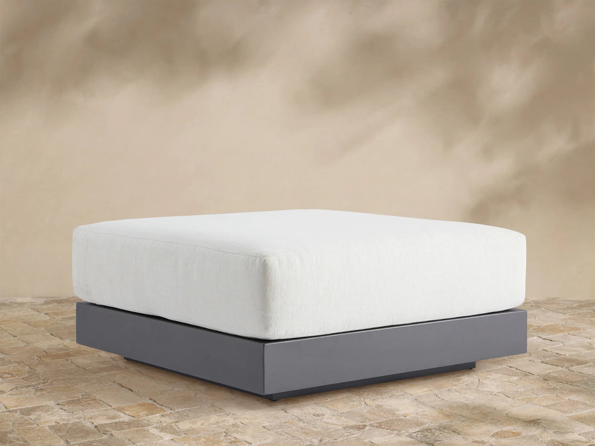 Naxos Outdoor Ottoman: rectangular white cushion on a dark gray aluminum base, centered on a light-colored patio.