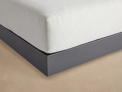 Naxos Outdoor Ottoman, square, with a thick white cushion atop a dark gray platform base, on a light patio floor.