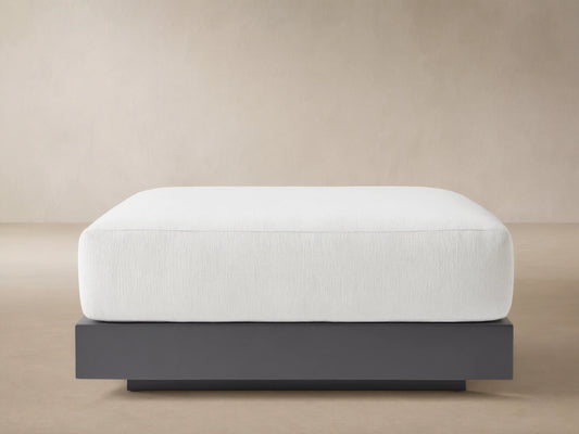Naxos Outdoor Ottoman: rectangular white cushion on a dark gray aluminum base, centered on a light-colored patio.