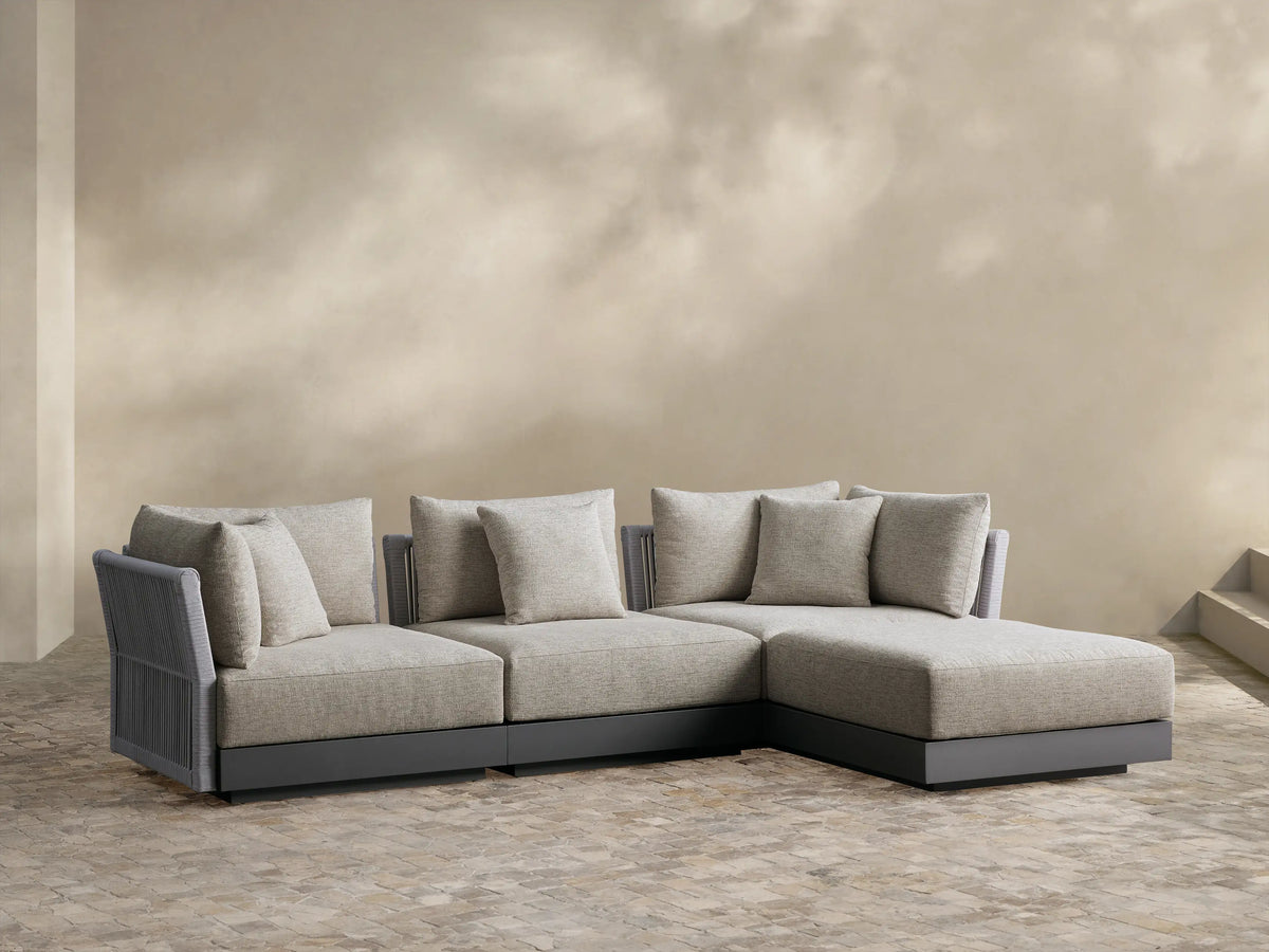 Naxos outdoor four-piece sectional: light beige fabric cushions, gray textured armrests, charcoal base, L-shaped on a paver patio.