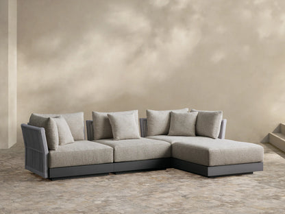 Naxos outdoor four-piece sectional: light beige fabric cushions, gray textured armrests, charcoal base, L-shaped on a paver patio.