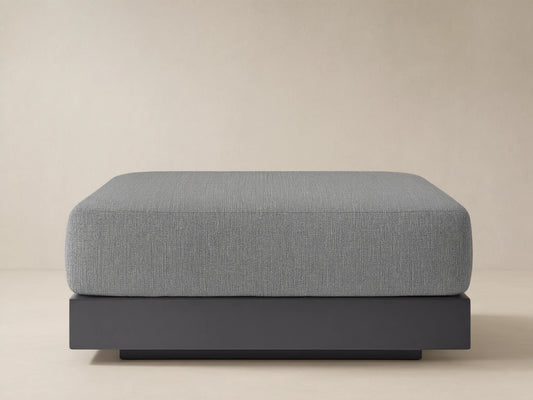 Naxos Outdoor Ottoman, rectangular grey upholstered cushion on a dark grey base, centered on a stone patio with light background.