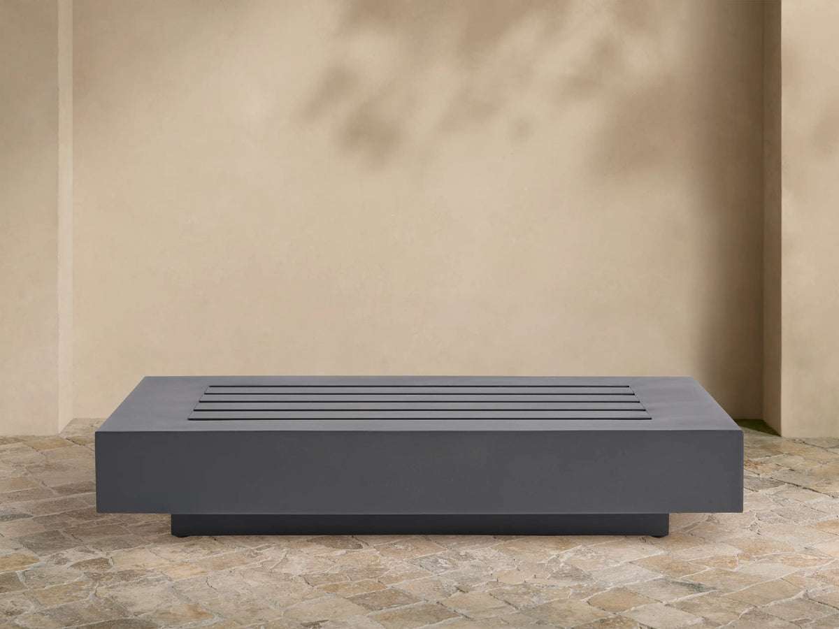 Rectangular Naxos Outdoor End Table in dark gray, featuring a modern slatted top, on a stone patio by a beige wall.