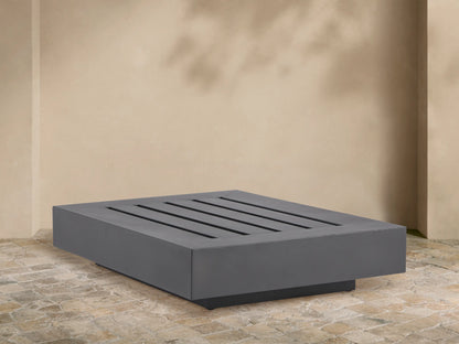 Rectangular Naxos Outdoor End Table in dark gray, featuring a modern slatted top, on a stone patio by a beige wall.