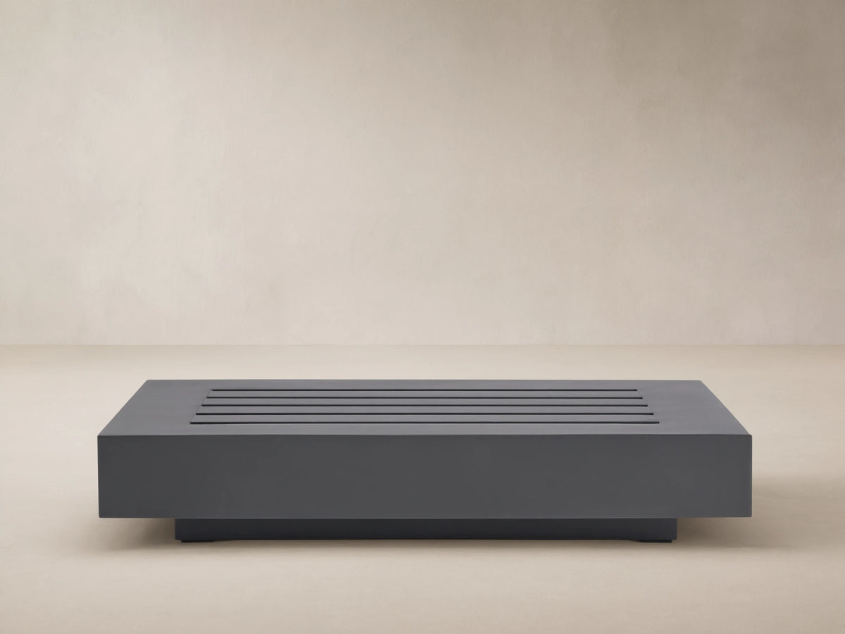 Rectangular Naxos Outdoor End Table in dark gray, featuring a modern slatted top, on a stone patio by a beige wall.