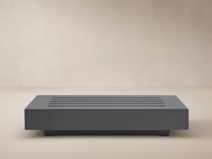 Rectangular Naxos Outdoor End Table in dark gray, featuring a modern slatted top, on a stone patio by a beige wall.