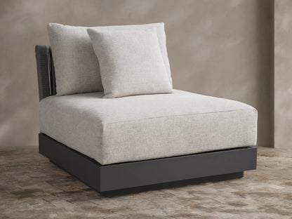 Naxos Outdoor Armless Chair with light grey textured cushions, dark charcoal base, and woven back, on a stone patio.