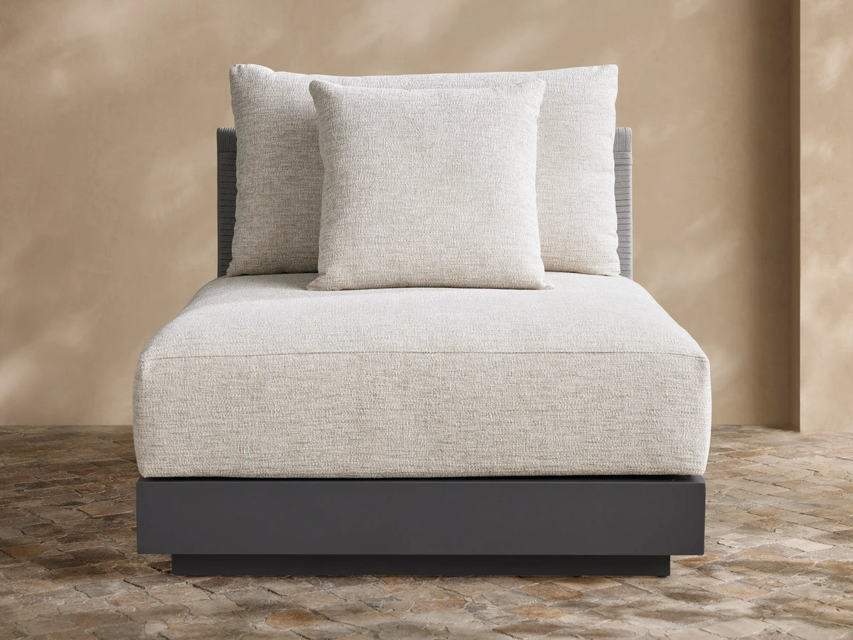 Naxos Outdoor Armless Chair with light grey textured cushions, dark charcoal base, and woven back, on a stone patio.