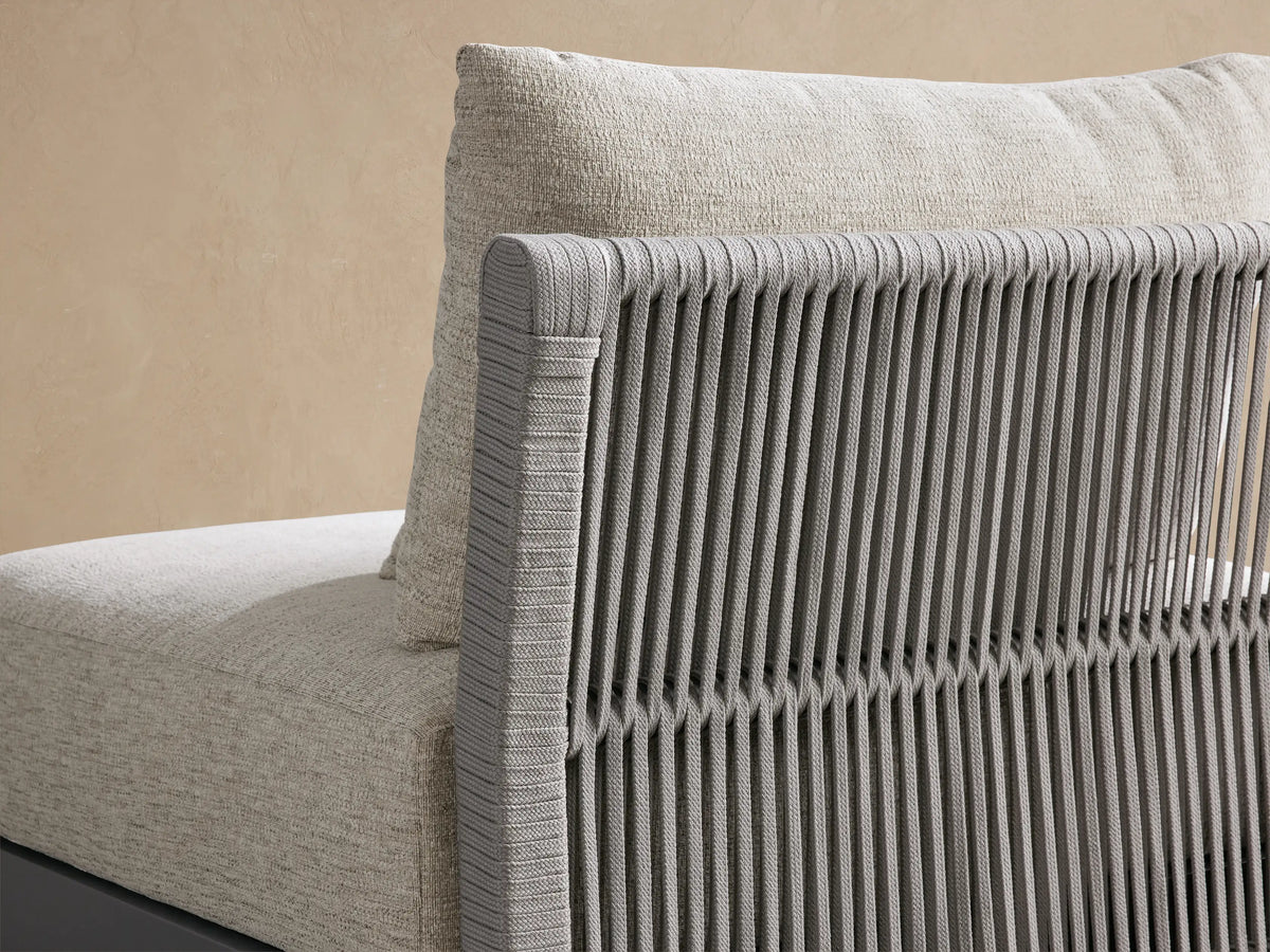 Naxos Outdoor Armless Chair with light beige textured cushions, dark gray frame & slatted back, on a stone floor.