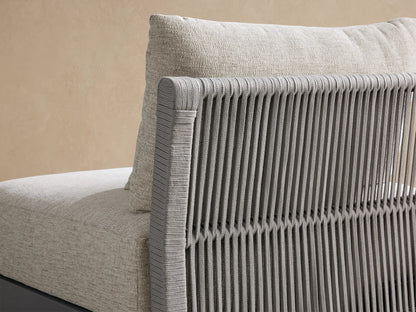 Naxos Outdoor Armless Chair with light beige textured cushions, dark gray frame & slatted back, on a stone floor.