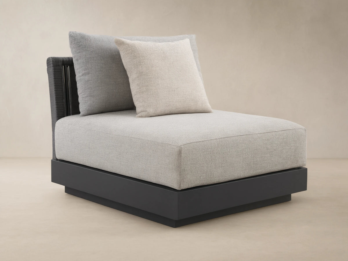 Naxos Outdoor Armless Chair with light grey textured cushions, dark charcoal base, and woven back, on a stone patio.