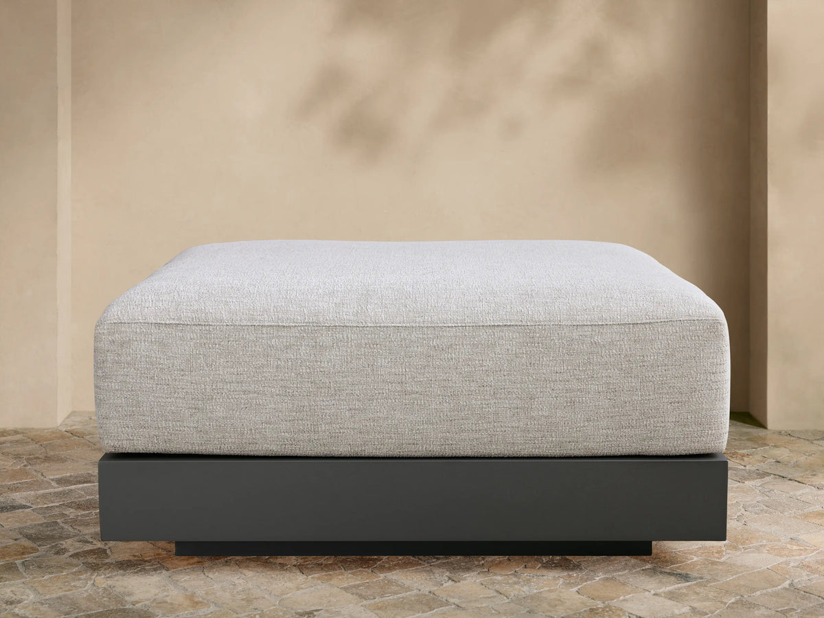 Naxos Outdoor Ottoman: light grey textured cushion on a dark grey rectangular base, centered on a tiled patio against a beige wall.