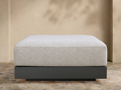 Naxos Outdoor Ottoman: light grey textured cushion on a dark grey rectangular base, centered on a tiled patio against a beige wall.