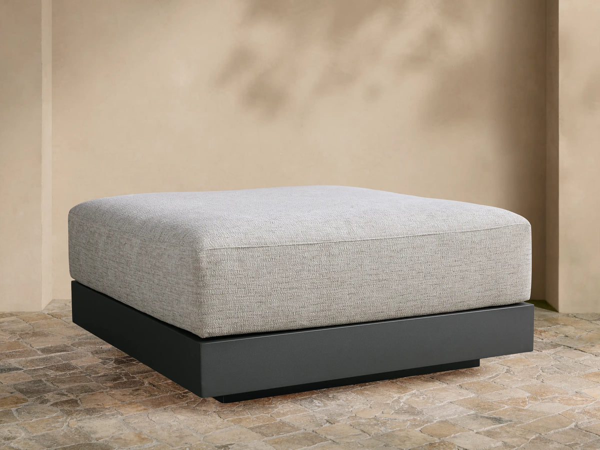 Naxos Outdoor Ottoman: light grey textured cushion on a dark grey rectangular base, centered on a tiled patio against a beige wall.
