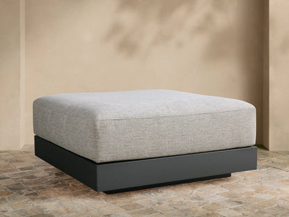 Naxos Outdoor Ottoman: light grey textured cushion on a dark grey rectangular base, centered on a tiled patio against a beige wall.
