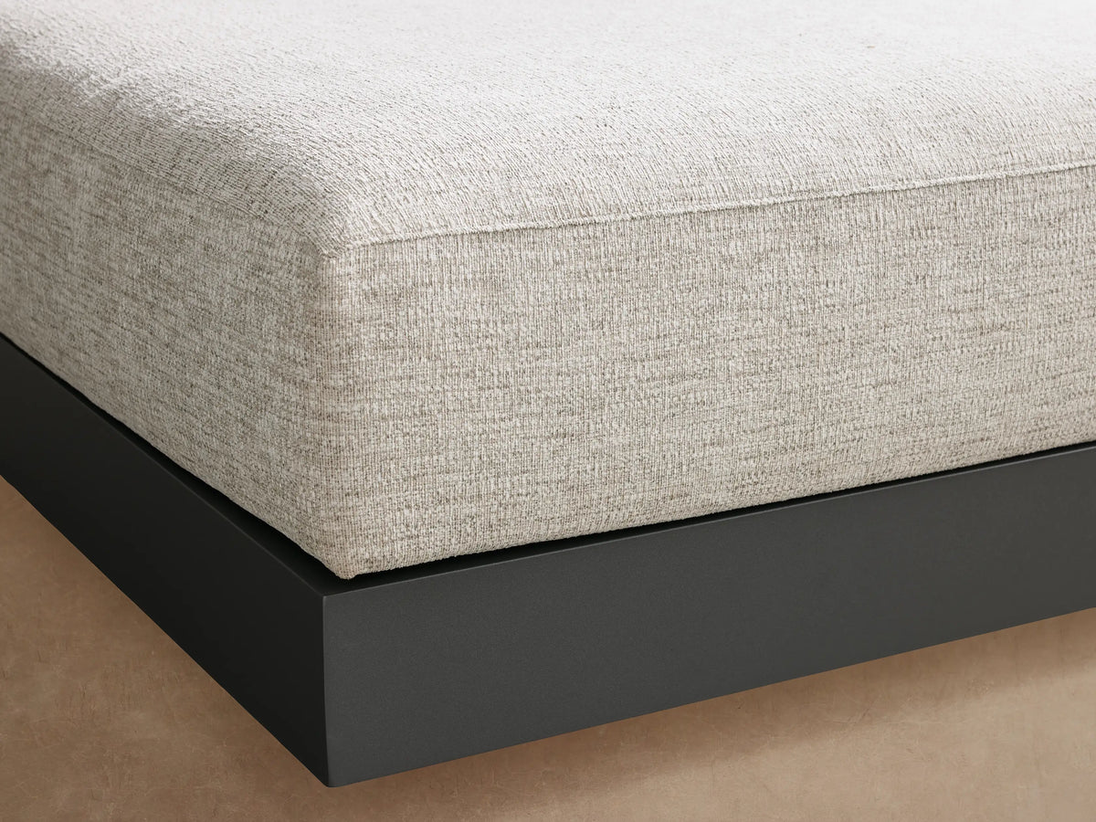 A square Naxos Outdoor Ottoman with a light gray textured cushion on a dark gray aluminum base, positioned on a stone patio.