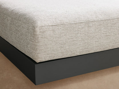 A square Naxos Outdoor Ottoman with a light gray textured cushion on a dark gray aluminum base, positioned on a stone patio.