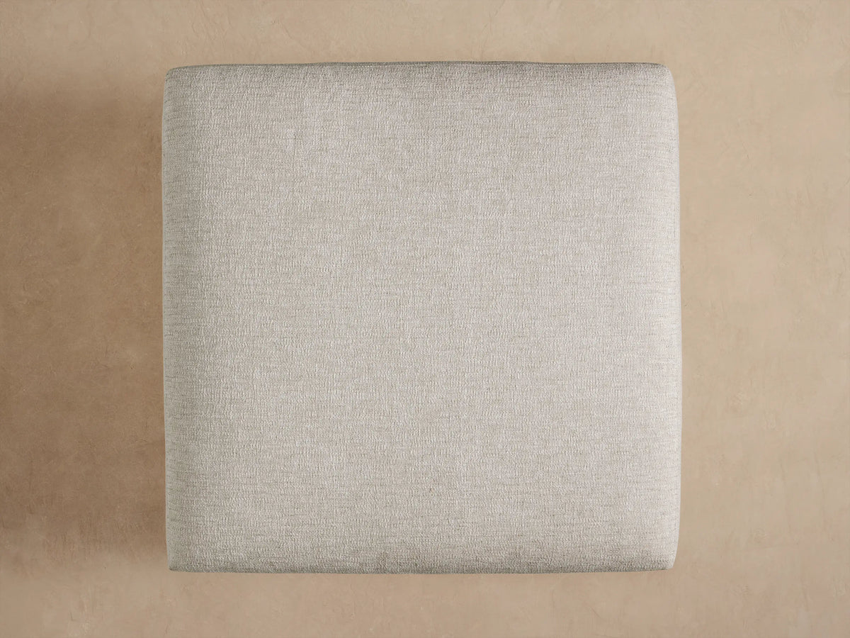 Close-up of Naxos Outdoor Ottoman's light beige textured cushion resting on a dark gray matte base, angled from top.