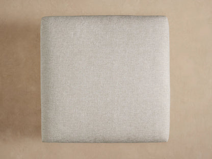 Close-up of Naxos Outdoor Ottoman's light beige textured cushion resting on a dark gray matte base, angled from top.