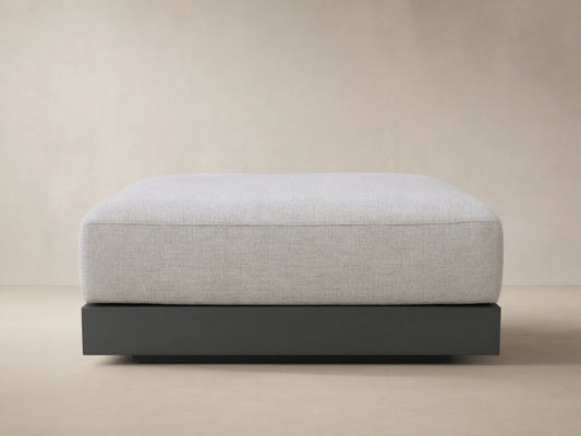 Naxos Outdoor Ottoman: light grey textured cushion on a dark grey rectangular base, centered on a tiled patio against a beige wall.