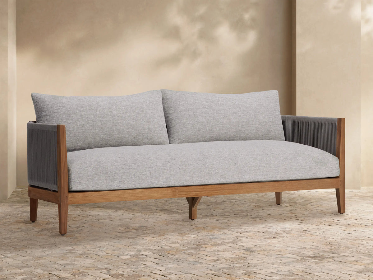 Newport Outdoor Sofa, front view. Natural teak wood frame, light grey woven cushions, dark grey woven arms, on stone patio.