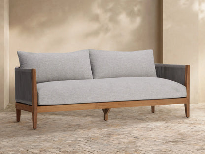 Newport Outdoor Sofa, front view. Natural teak wood frame, light grey woven cushions, dark grey woven arms, on stone patio.