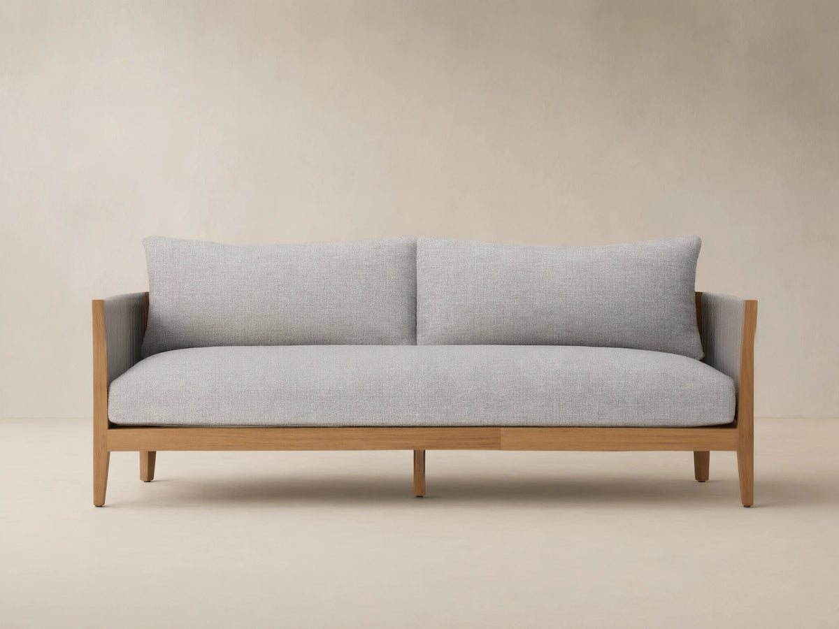 Newport Outdoor Sofa, front view. Natural teak wood frame, light grey woven cushions, dark grey woven arms, on stone patio.