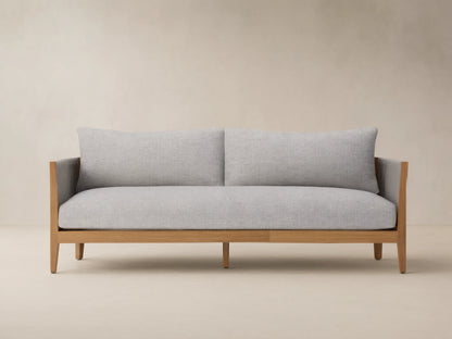 Newport Outdoor Sofa, front view. Natural teak wood frame, light grey woven cushions, dark grey woven arms, on stone patio.