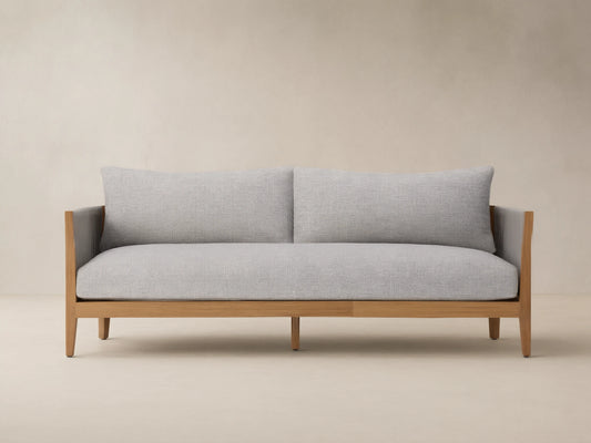 Newport Outdoor Sofa, front view. Natural teak wood frame, light grey woven cushions, dark grey woven arms, on stone patio.