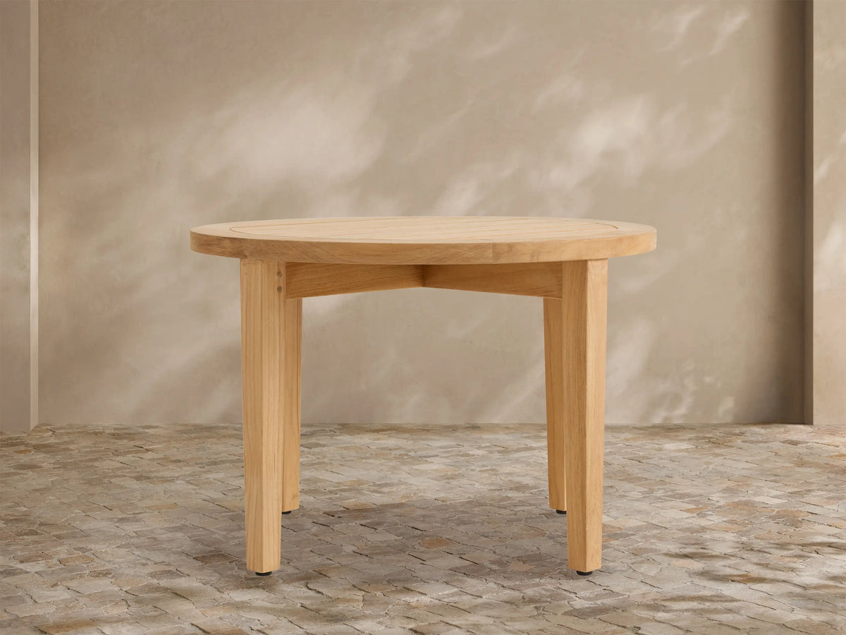 Newport Outdoor Coffee Table. Round, light-toned teak wood with a slatted top and four sturdy legs on a stone patio.
