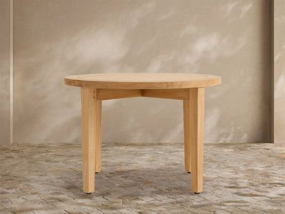 Newport Outdoor Coffee Table. Round, light-toned teak wood with a slatted top and four sturdy legs on a stone patio.