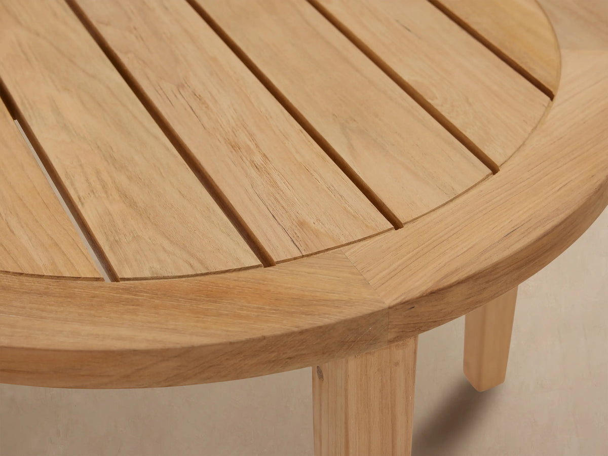 Newport Outdoor Coffee Table: round, light-colored natural teak wood, slatted top, sturdy legs on a textured patio against a beige wall.