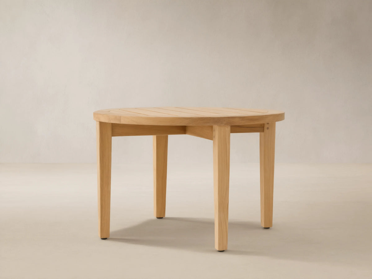 Newport Outdoor Coffee Table. Round, light-toned teak wood with a slatted top and four sturdy legs on a stone patio.