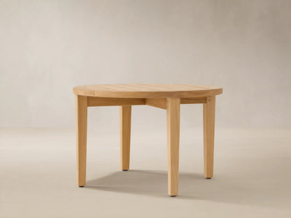 Newport Outdoor Coffee Table. Round, light-toned teak wood with a slatted top and four sturdy legs on a stone patio.