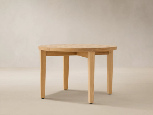 Newport Outdoor Coffee Table. Round, light-toned teak wood with a slatted top and four sturdy legs on a stone patio.