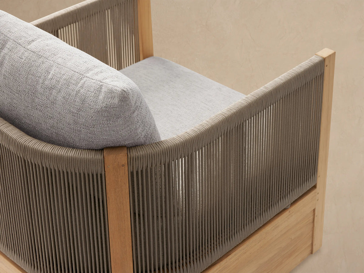Front view of Newport Outdoor Lounge Chair with natural teak frame, light grey textured cushions, and woven armrests.