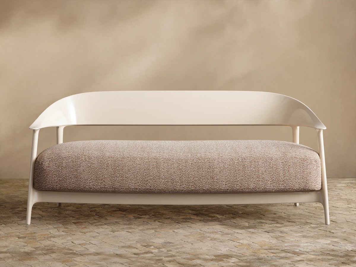 Nia Outdoor Sofa. Cream frame, curved backrest, light brown speckled cushion, against a beige wall & tiled floor.