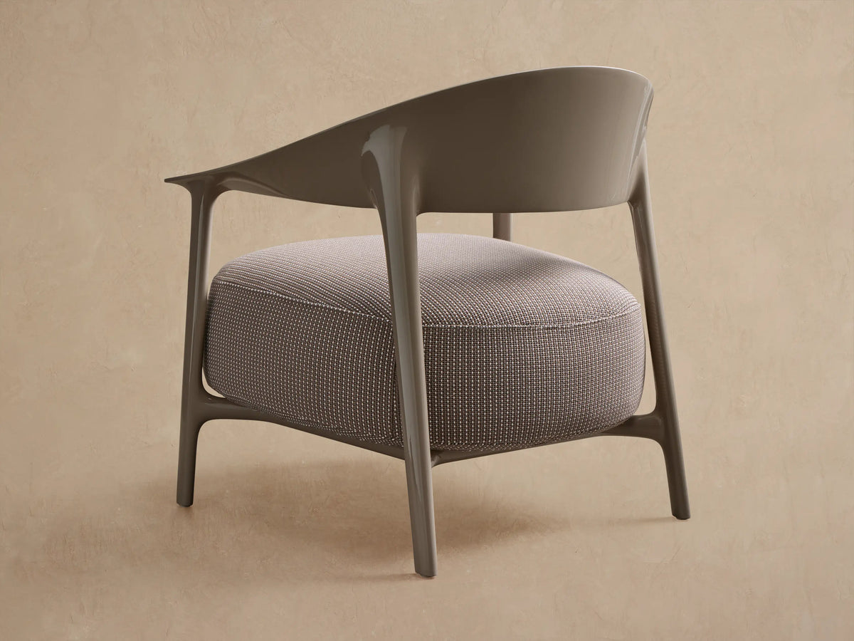 Nia Outdoor Lounge Chair. Angled view of a light taupe, glossy frame with a curved backrest & round, textured light beige cushion.