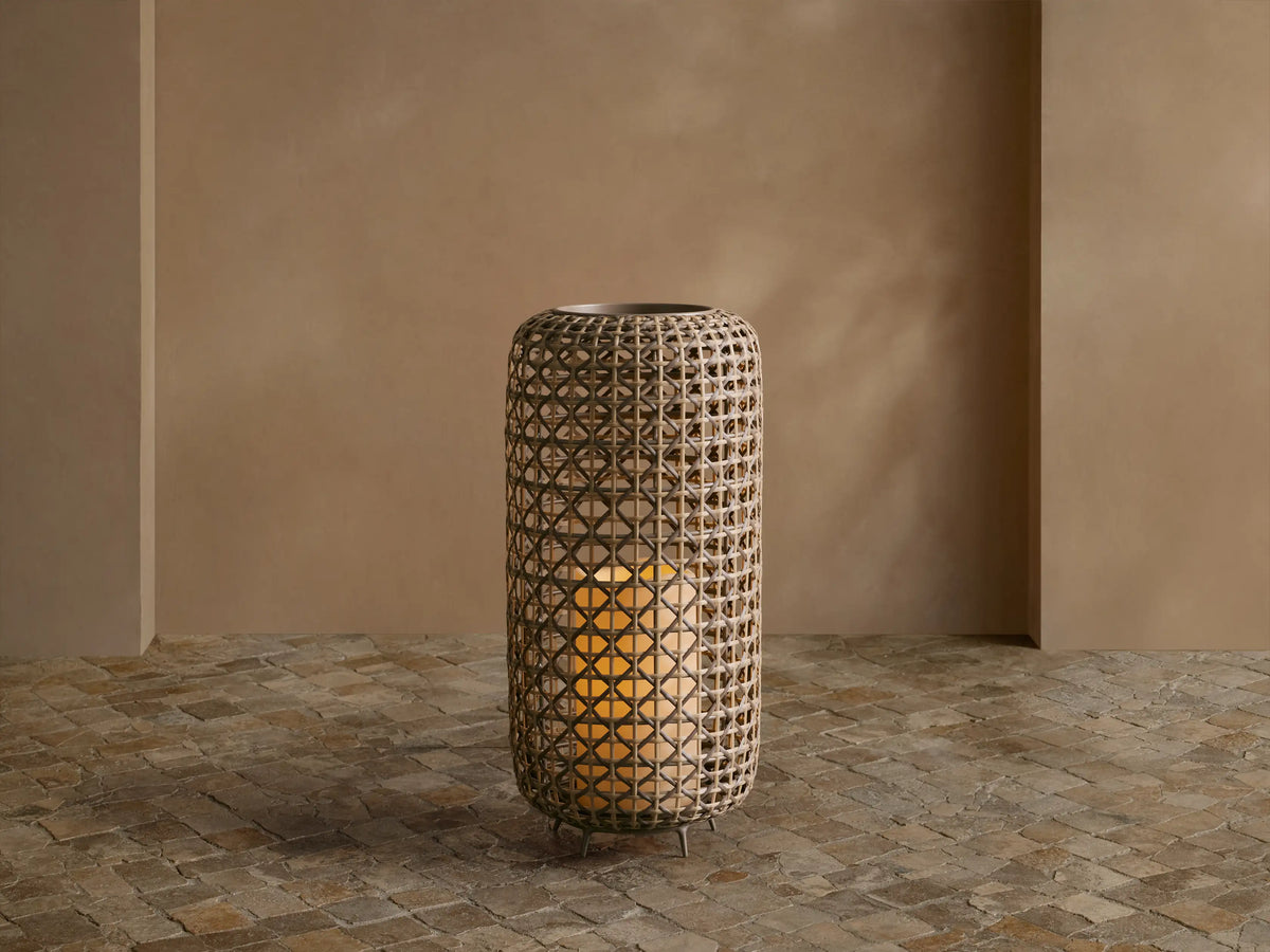 Noale Outdoor Lantern, tall cylindrical handwoven wicker with warm glow from inside, standing on a stone floor.