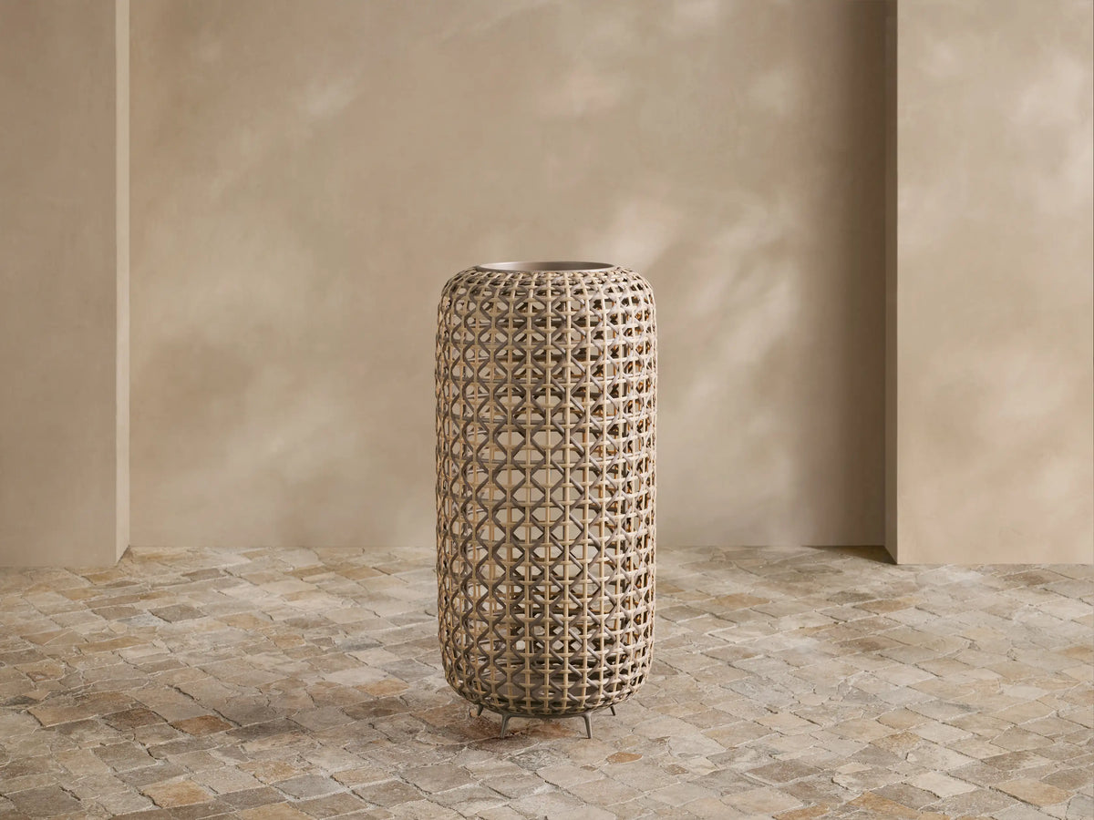 Noale Outdoor Lantern, tall cylindrical handwoven wicker with warm glow from inside, standing on a stone floor.