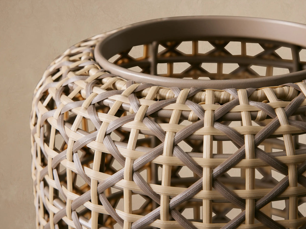 Tall, cylindrical Noale Outdoor Lantern in light brown woven wicker with a geometric pattern, standing on a textured stone floor.
