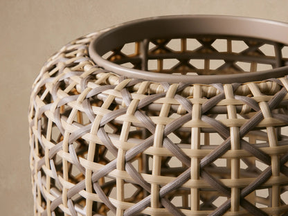 Tall, cylindrical Noale Outdoor Lantern in light brown woven wicker with a geometric pattern, standing on a textured stone floor.
