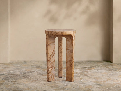 Odessa Outdoor End Table. Cylindrical top, three thick, curved legs, light tan with dark brown stone-like veining, on tiled floor.